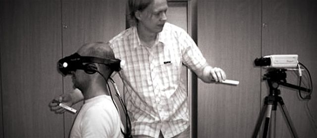 One person sitting with a VR set, while another pokes at a camera